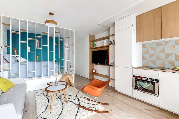 Cucina lineare laccata bianca con pensili in essenza collegata con parete soggiorno, in un piccolo monolocale con elemento divisorio freestanding tra divano e letto.